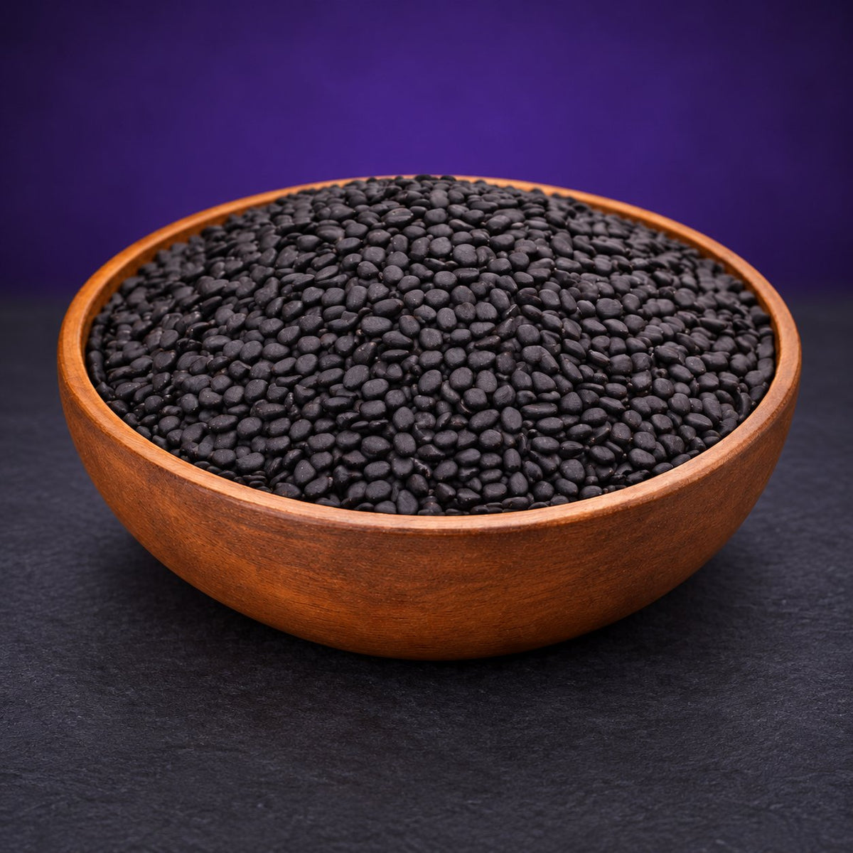 Black sesame seeds in a wooden bowl on a matte charcoal and subtle royal-purple background.