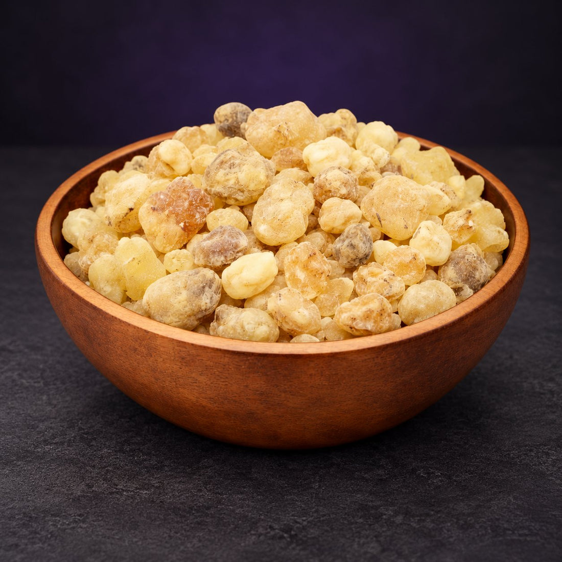 Pale golden frankincense resin pieces in a wooden bowl against a dark purple and charcoal background.